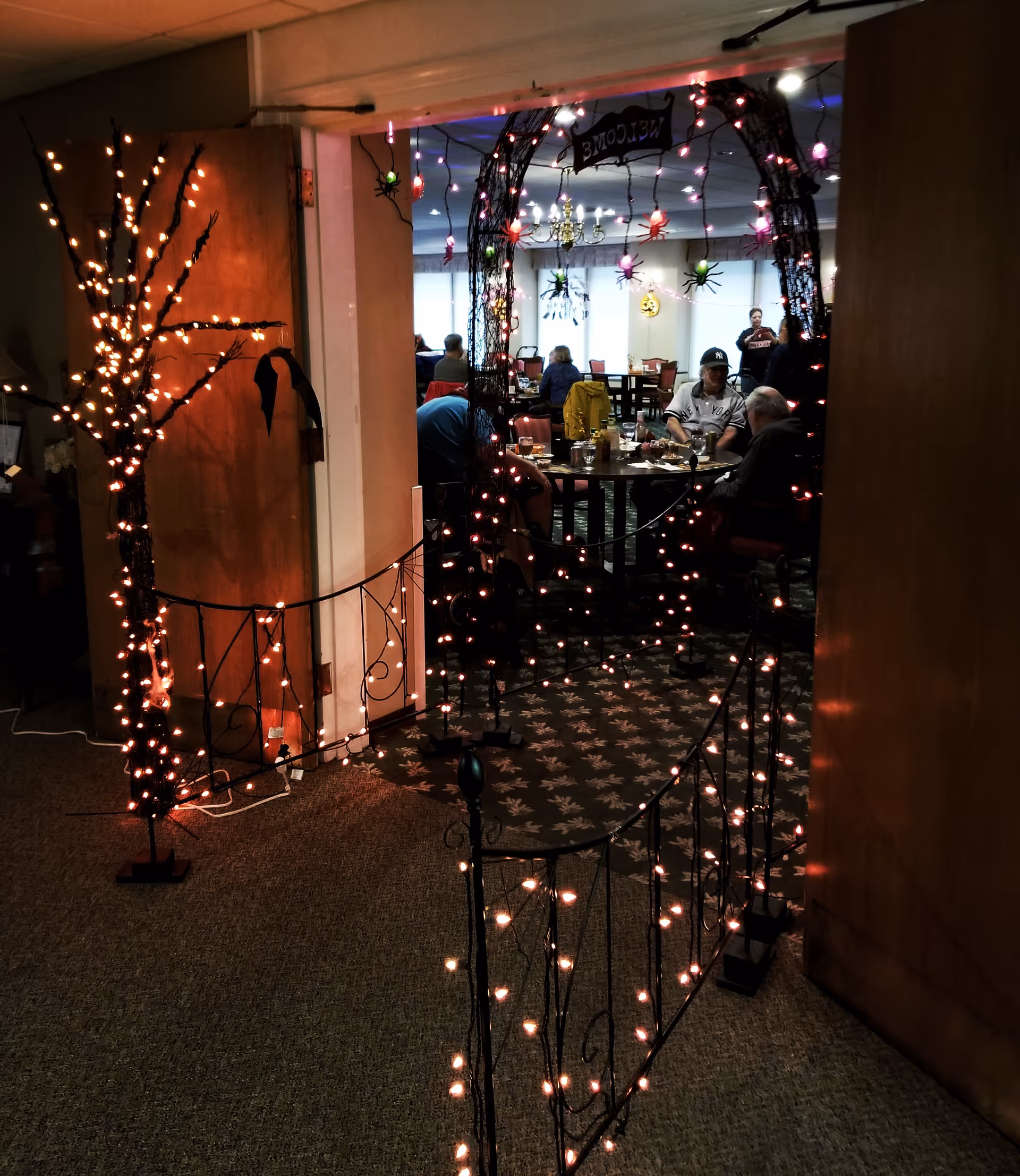 Interior view of a dining area decorated with Halloween-themed lights and ornaments, including a lighted archway with hanging spiders and a 'Welcome' sign. Several people are seated at tables eating and socializing in the background.