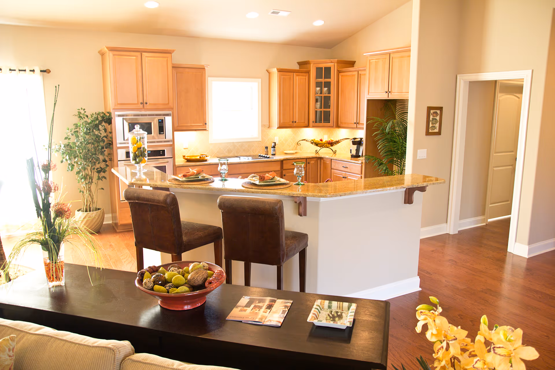 Bright and spacious kitchen with wooden cabinets, a granite countertop breakfast bar with two brown leather chairs, and various decorative plants and items. The kitchen features built-in microwave and oven, a window above the sink, and warm wooden flooring.