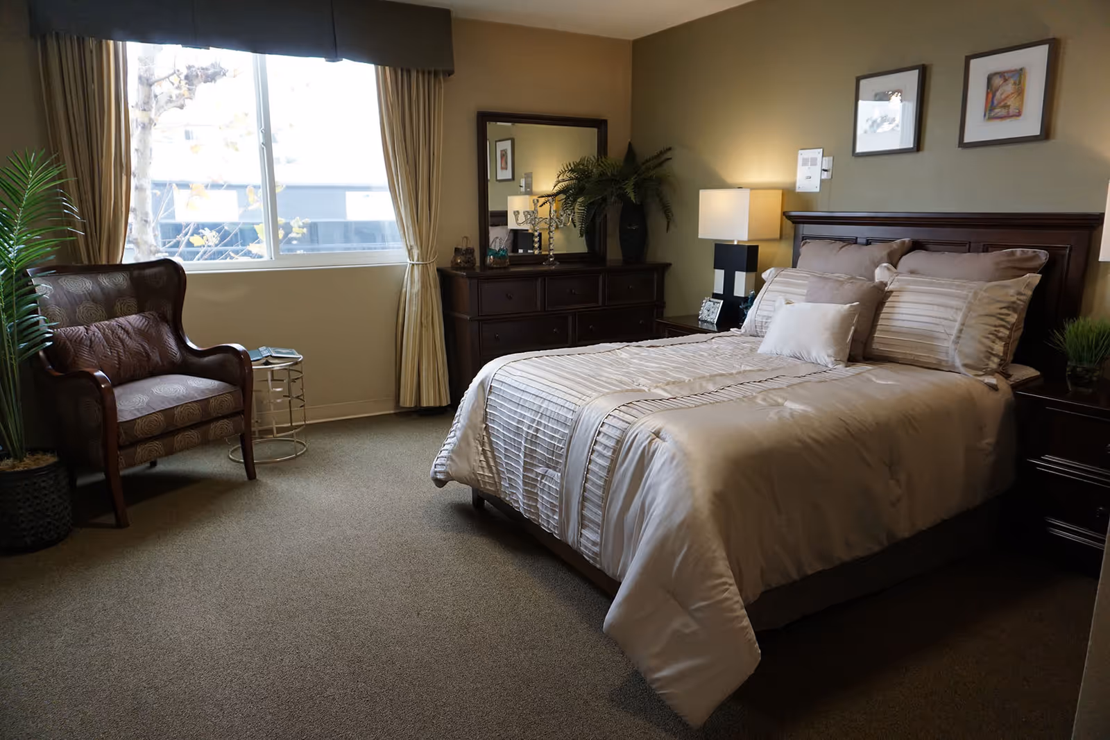 A cozy bedroom with a neatly made bed featuring beige and cream bedding. There is a wooden headboard, two framed pictures on the wall above the bed, and a bedside table with a lamp and a small plant. Across from the bed is a wooden dresser with a large mirror, decorative items, and a lamp. A comfortable armchair and a small round side table are positioned near a window with beige curtains. A potted plant is placed beside the armchair.