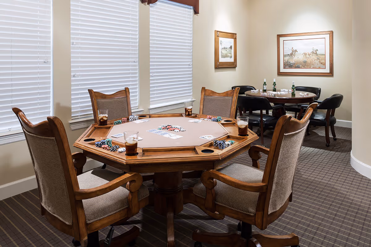 A cozy game room with two card tables set up for poker. The foreground table is octagonal with wooden chairs and poker chips, cards, and drinks on it. The background table is round with black chairs, poker chips, cards, and drinks. The room has beige walls, carpeted floor, and framed artwork on the walls.