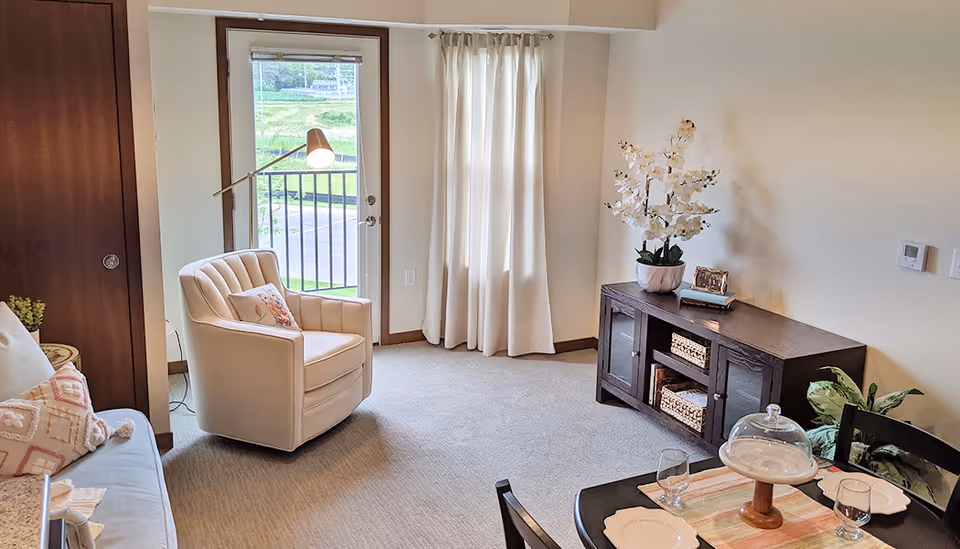 A cozy living room area in a senior living facility with a cream-colored armchair, a floor lamp, a wooden cabinet with decorative items including a white orchid plant, and a small dining table set with plates, glasses, and a covered cake stand. There is a door with a window leading outside and light-colored curtains.