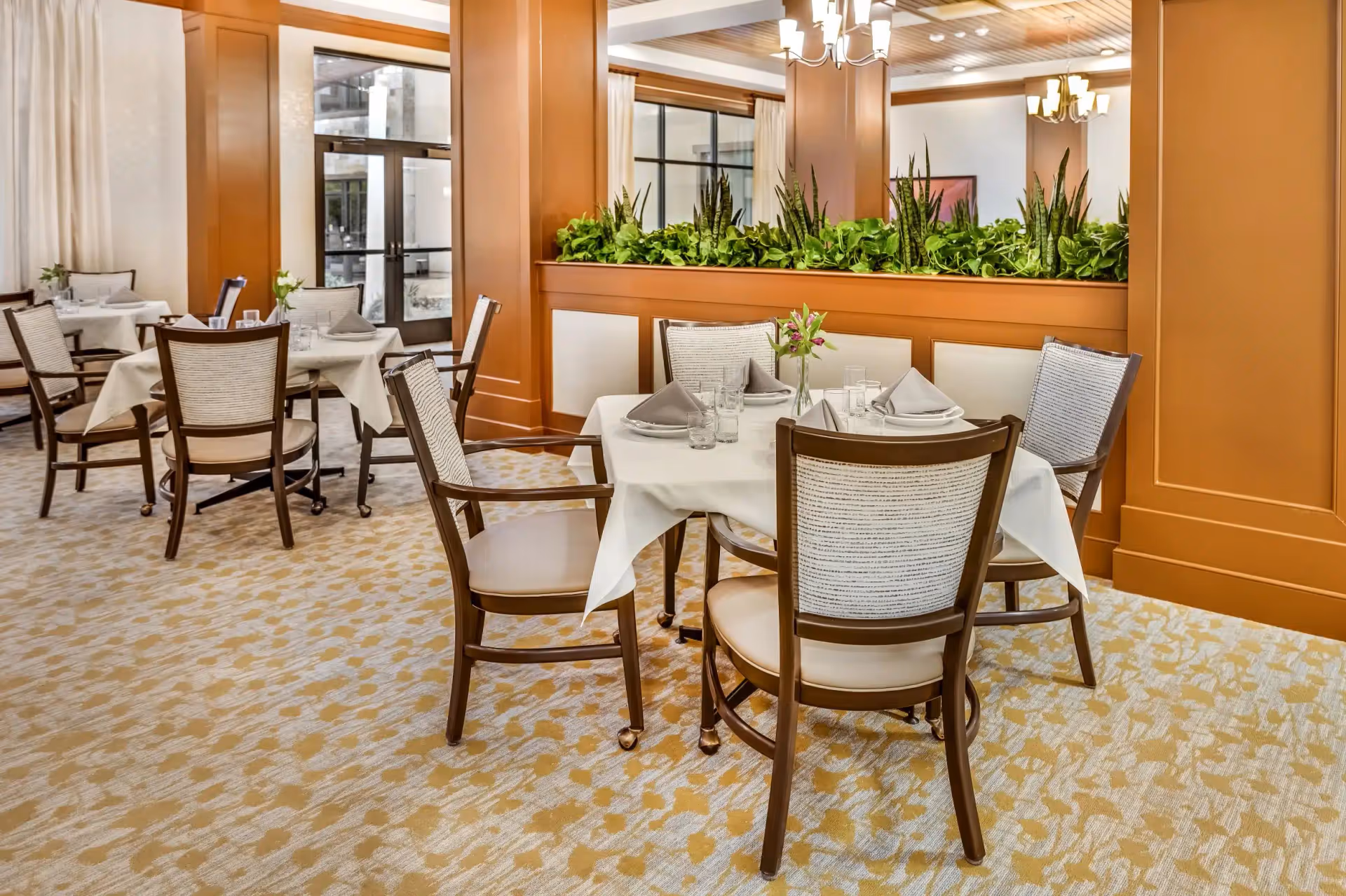 Well-lit dining room with round tables set with napkins and glasses, wooden chairs, and a central planter divider.