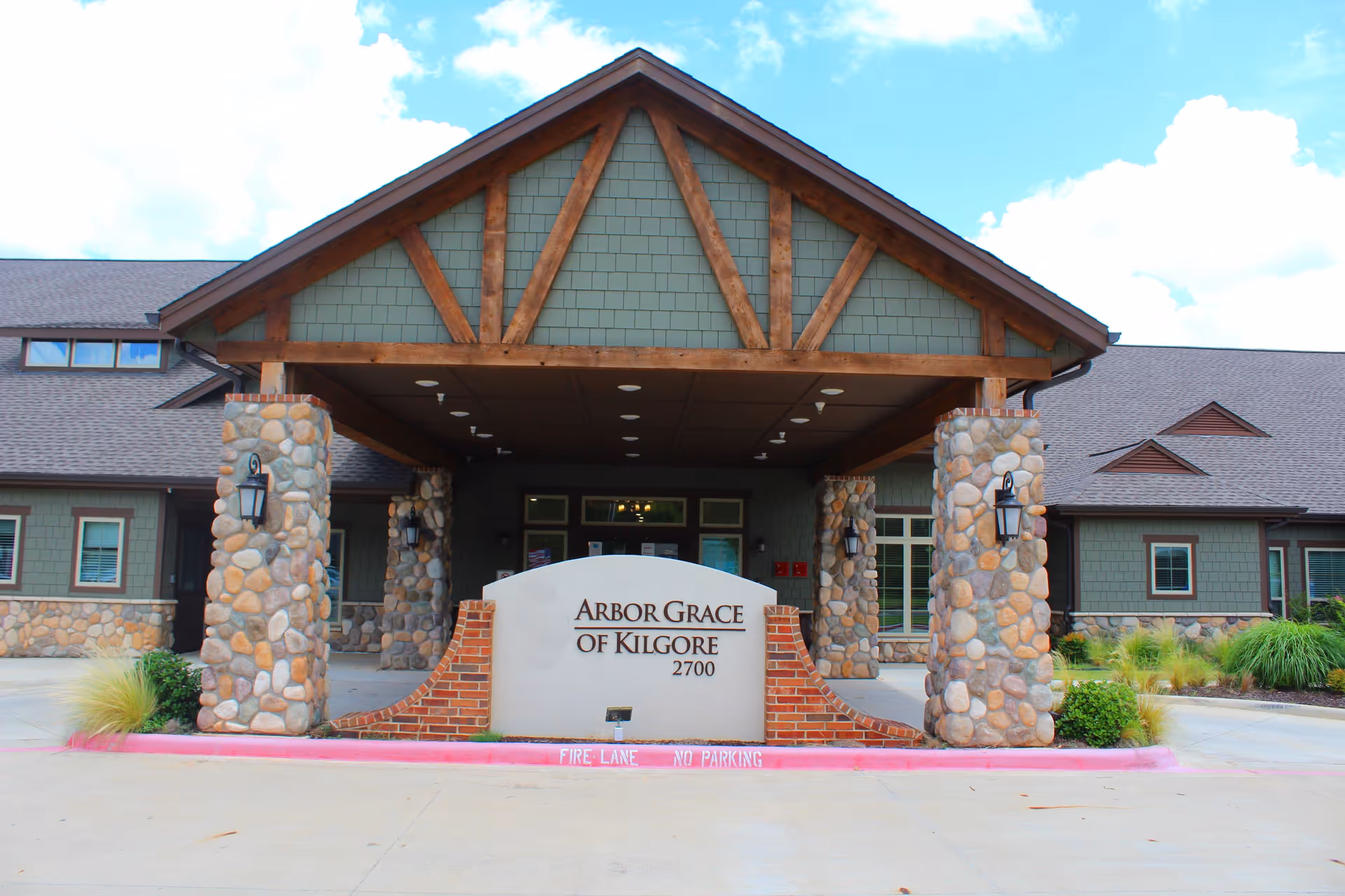 Front exterior view of Arbor Grace of Kilgore facility showing a covered entrance supported by stone pillars, with a sign displaying the facility name and address 2700. The building has green siding with stone accents and a brown roof under a partly cloudy sky.