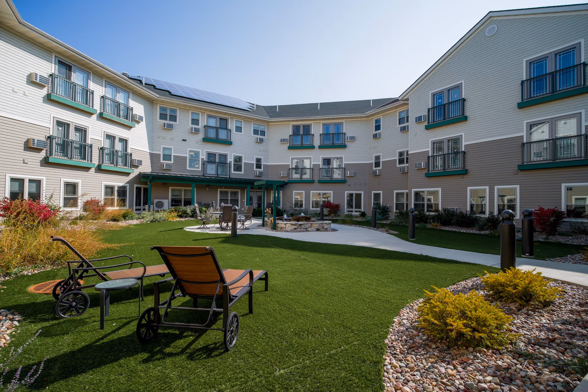 Outdoor courtyard area of a senior living facility with green artificial grass, lounge chairs, a small table, a fire pit surrounded by chairs, and a three-story building with balconies and windows in the background under a clear blue sky.