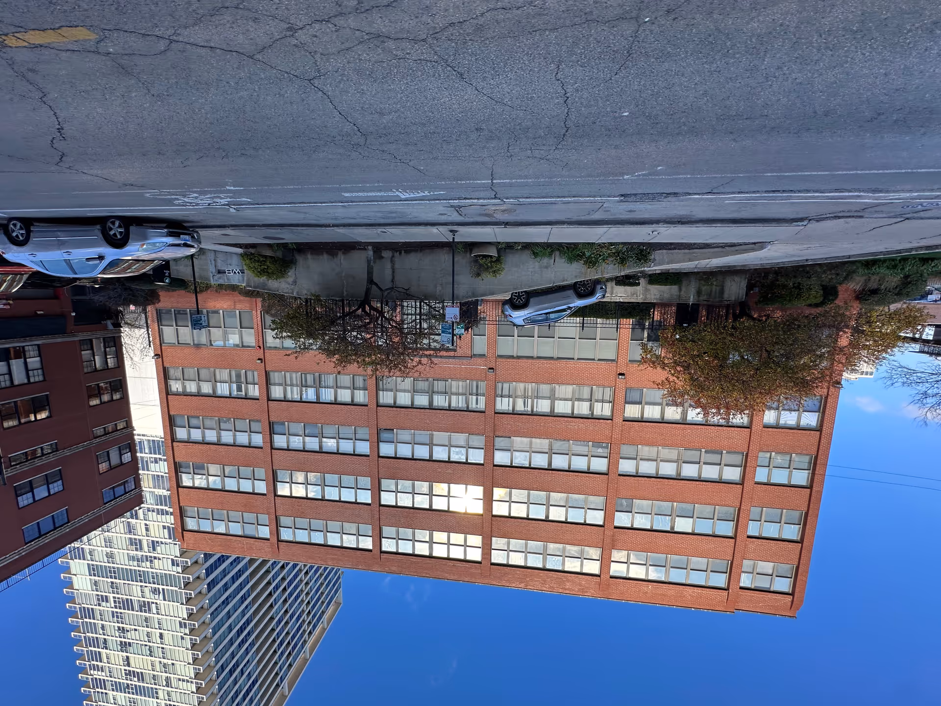 Exterior view of a multi-story red brick building with many windows, a concrete ramp with a silver SUV parked on it, some trees and bushes in front, and a clear blue sky above. There are other buildings visible in the background and a few cars parked along the street in front.