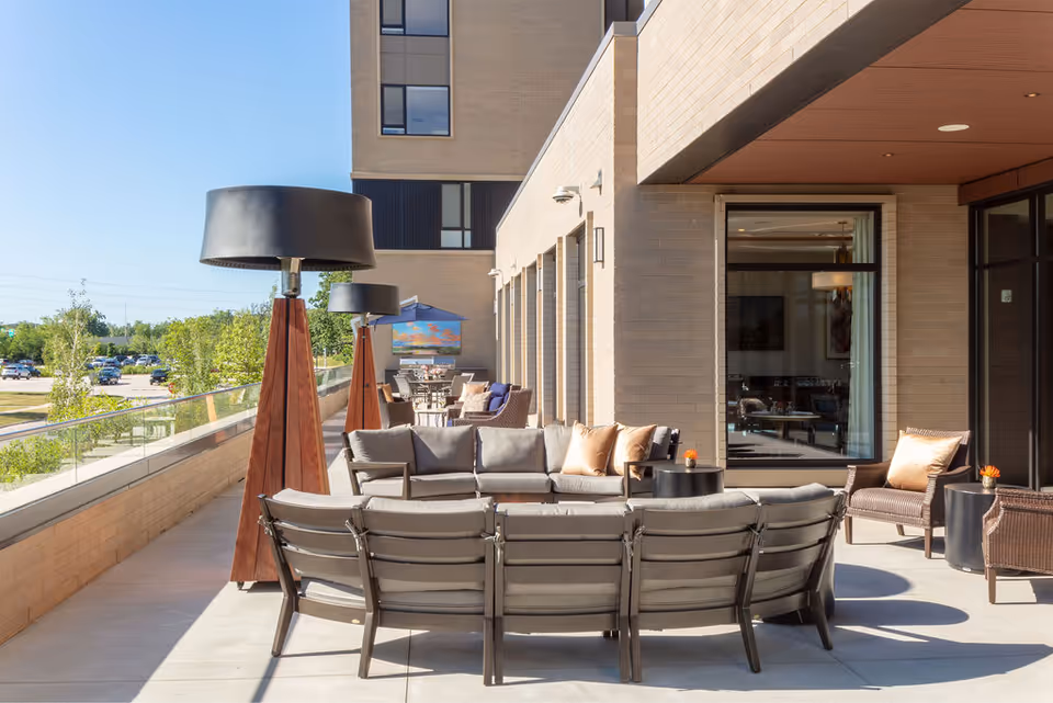 Outdoor terrace with a curved sofa, lounge chairs, tall floor lamps and patio tables beside a modern building.