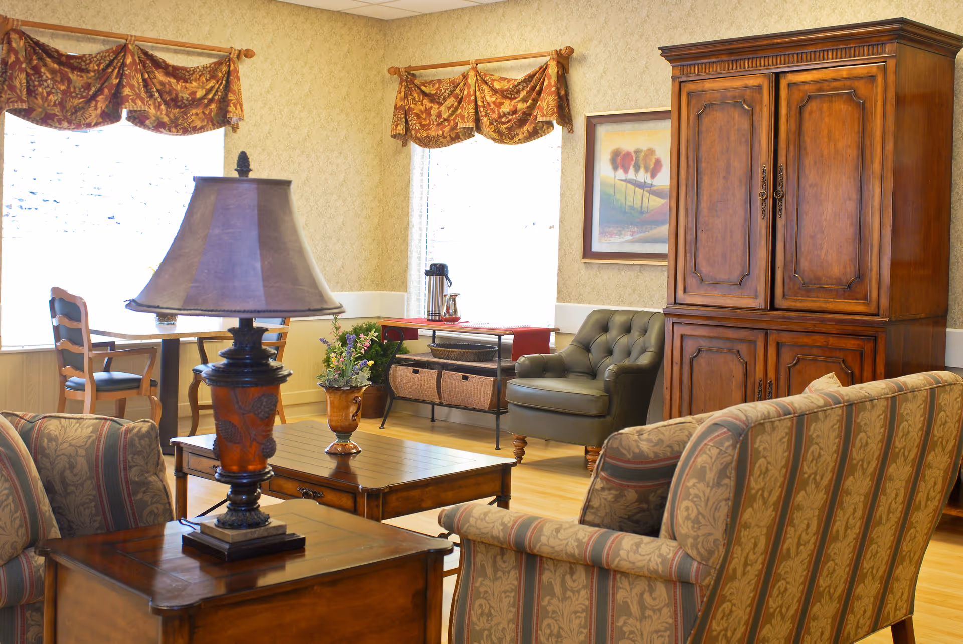 A cozy senior living room with patterned sofas, wooden coffee and side tables, a lamp, an armchair, and a large wooden armoire by bright windows.