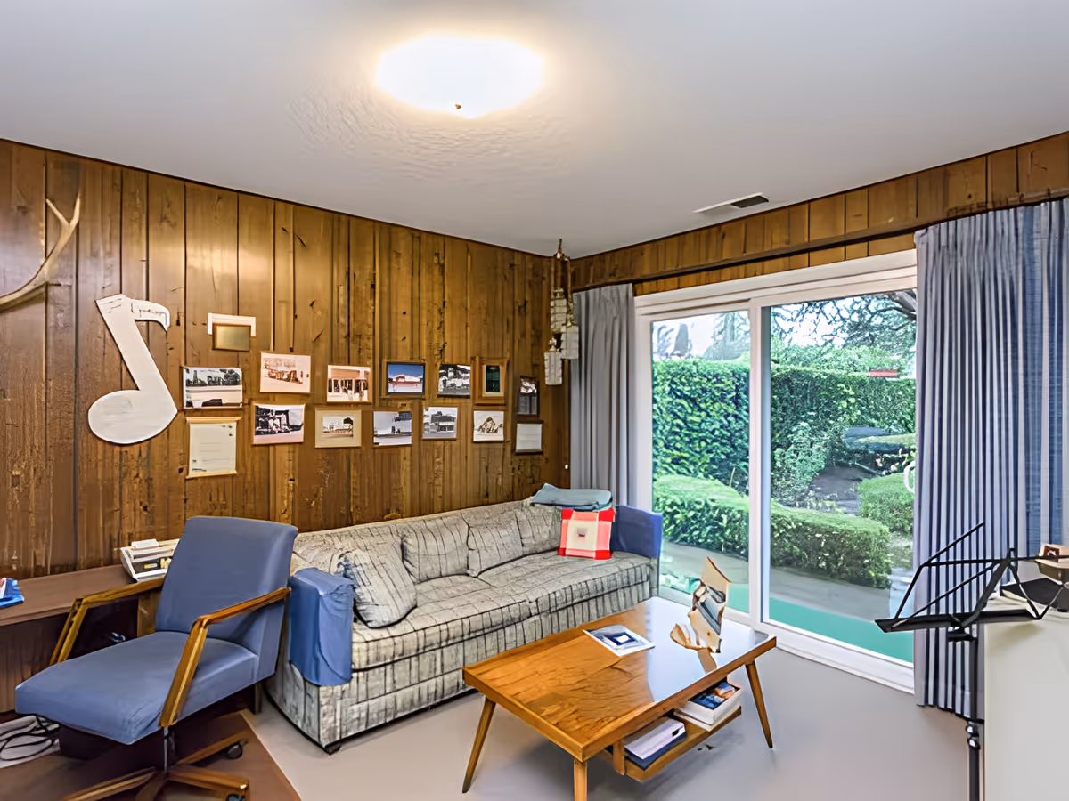 A cozy living room with wood-paneled walls, a patterned sofa with a red and white pillow, a blue armchair, a wooden coffee table with books and papers, and a large window with blue curtains showing a green garden outside. Various framed photos and a large musical note decoration hang on the wall.