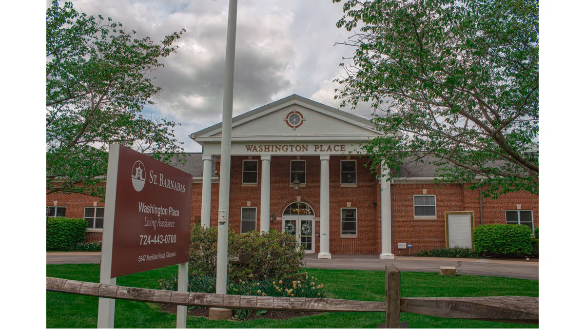 Front exterior view of the St. Barnabas Washington Place building, a brick structure with white columns and a sign in front displaying the facility name, contact number, and address. Trees and greenery surround the area under a cloudy sky.