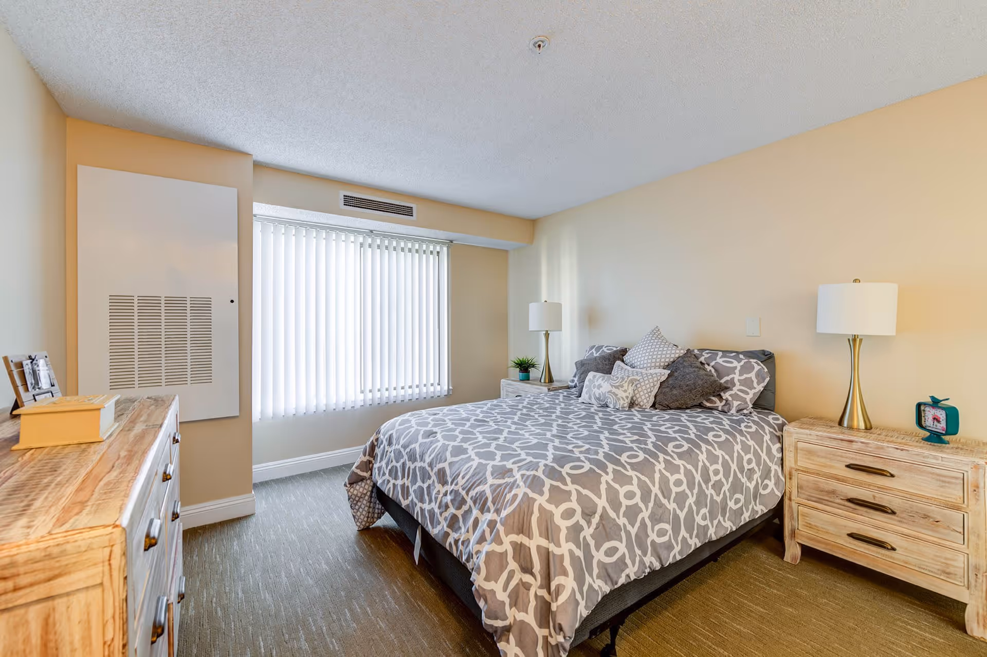 A bedroom with a large bed covered in a gray and white patterned comforter and multiple pillows. There are two wooden nightstands on either side of the bed, each with a lamp. A wooden dresser with a few decorative items is positioned against the opposite wall. A large window with vertical blinds allows natural light into the room. The walls are painted a soft beige color.