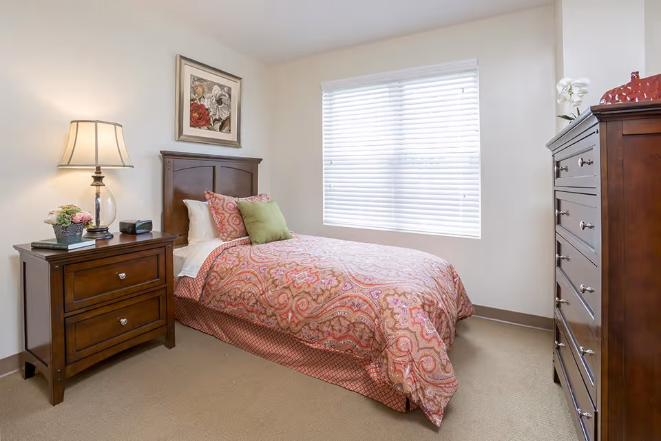 A cozy bedroom with a single bed covered in a patterned red and pink bedspread, two pillows including a green one, a wooden headboard, a wooden nightstand with a lamp, a flower pot, and a clock, a large window with white blinds, and a tall wooden dresser with a decorative item and a white orchid on top.