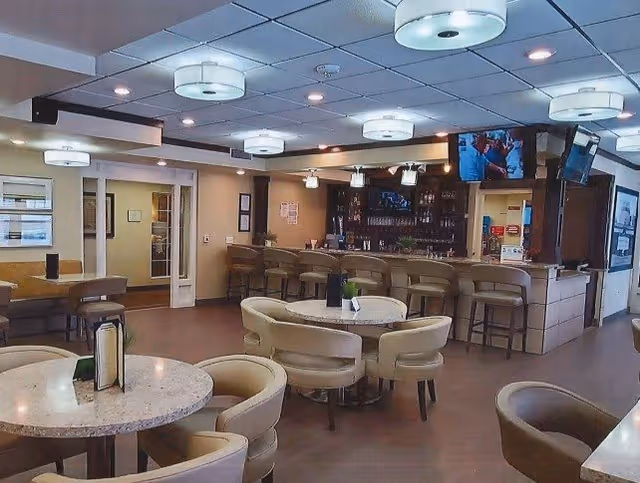 Interior view of a lounge area with round tables and cushioned chairs arranged around them. There is a bar counter with bar stools and multiple ceiling lights. Two televisions are mounted above the bar area, and the space has a warm, inviting atmosphere.