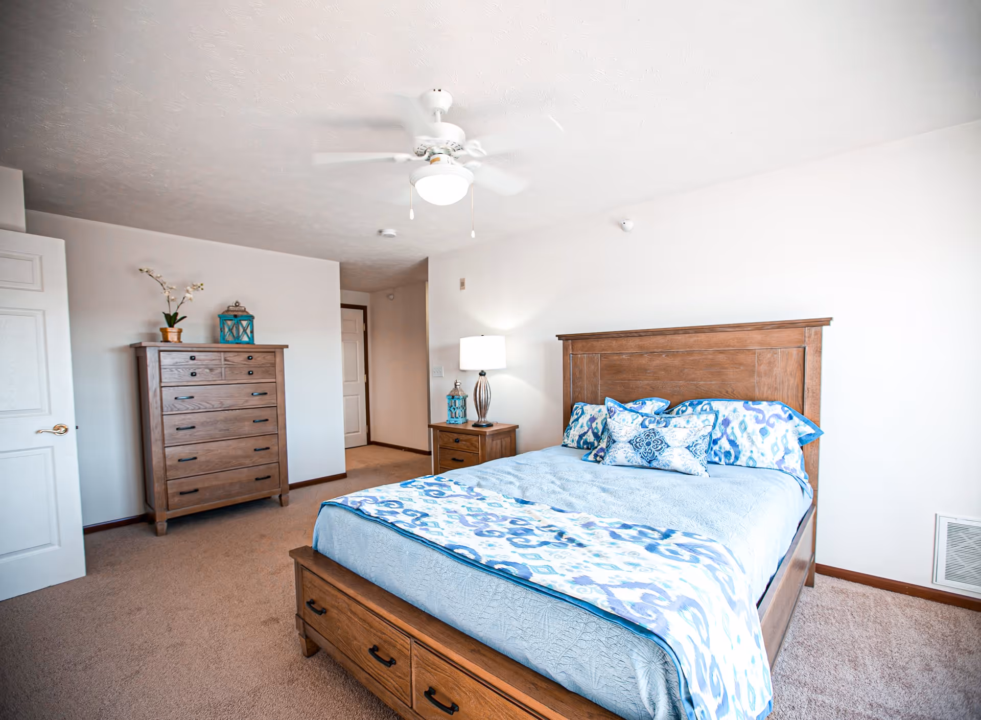 A bright bedroom with a wooden bed frame and matching dresser and nightstand. The bed is made with light blue bedding and patterned pillows. A ceiling fan with a light is mounted on the ceiling. The room has beige carpet and white walls with a door slightly open on the left side.