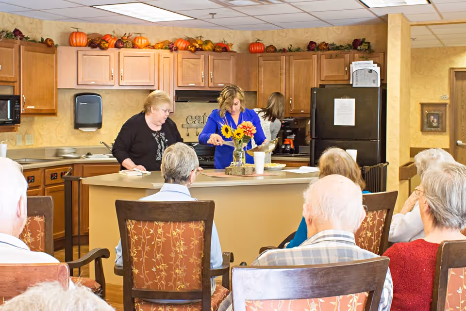 A group of elderly people seated in chairs facing a kitchen area where three women are preparing and serving food. The kitchen has wooden cabinets decorated with autumn-themed items like pumpkins and gourds. A vase with sunflowers is placed on the counter.
