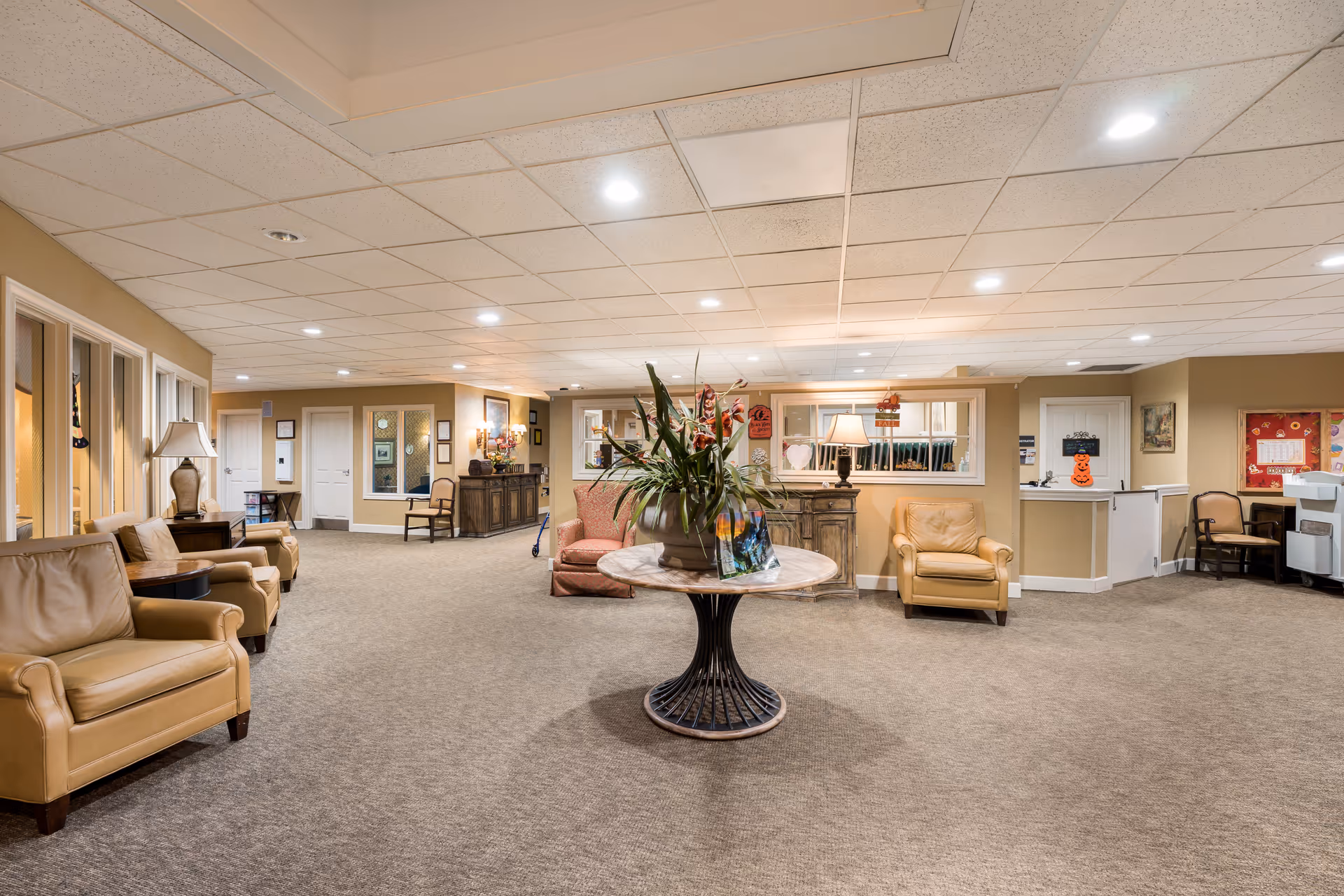 A spacious and well-lit senior living facility common area with beige walls and carpeted floors. The room features several comfortable tan leather armchairs arranged along the walls, a round table with a large floral arrangement in the center, and various pieces of furniture including lamps, chairs, and cabinets. The ceiling has recessed lighting and a drop ceiling design. There are doors and windows along the walls, and some seasonal decorations are visible.