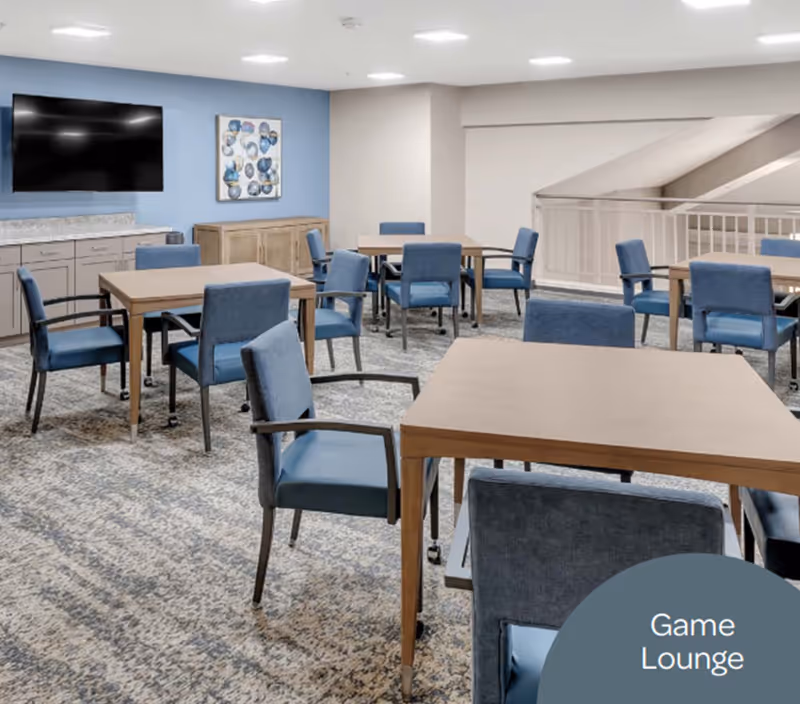 A bright game lounge with several wooden tables, blue upholstered chairs, a wall-mounted TV, and artwork on the wall.