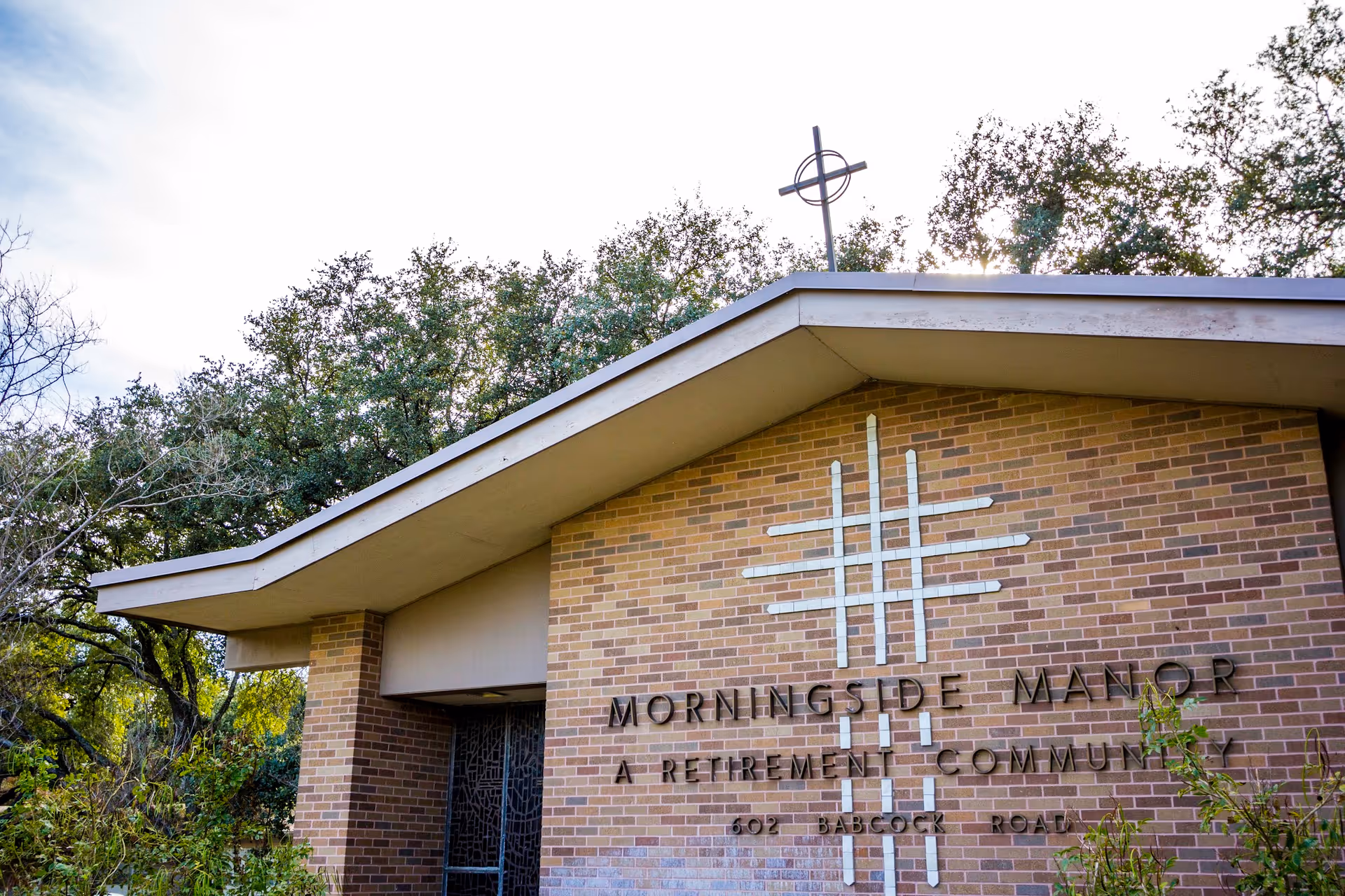 Brick facade of Morningside Manor retirement community entrance with a cross and signage under a sloped roof and trees in the background.