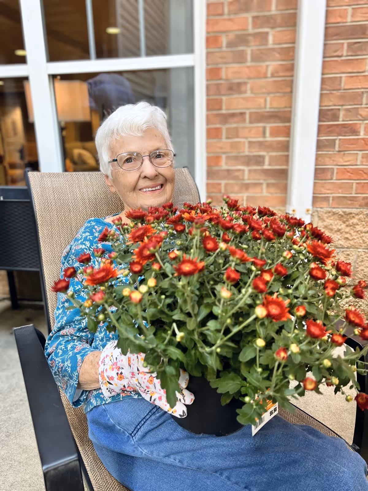 An elderly woman with white hair and glasses sitting outdoors in a chair, smiling and holding a large pot of red flowers. She is wearing a blue floral shirt, gardening gloves, and blue pants. The background shows a brick wall and a window.