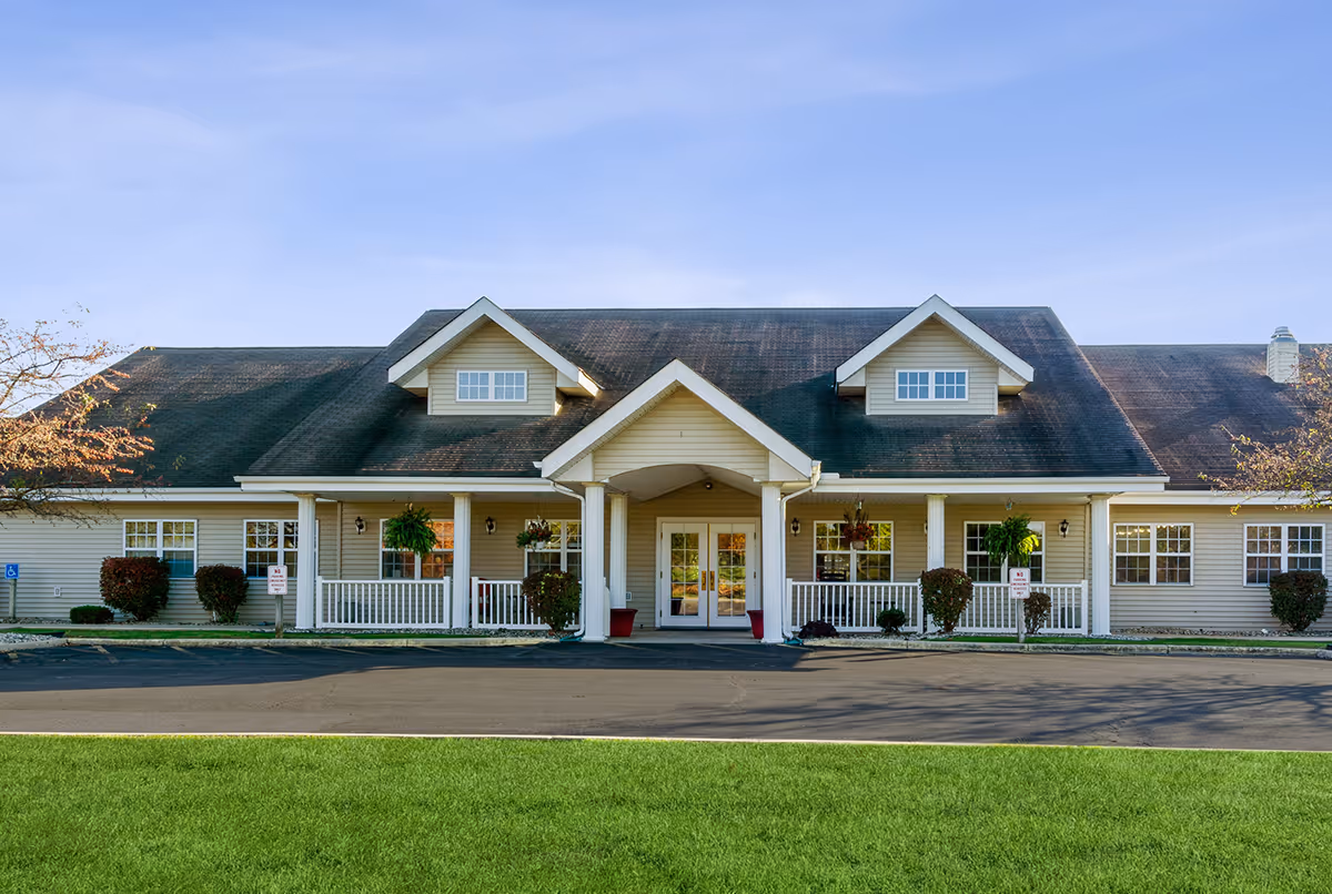 Front exterior view of a single-story building with a dark shingled roof, beige siding, white trim, and a covered entrance with columns. There are several windows, hanging plants, and shrubs along the front. The building is set against a clear blue sky with a green lawn and paved driveway in front.