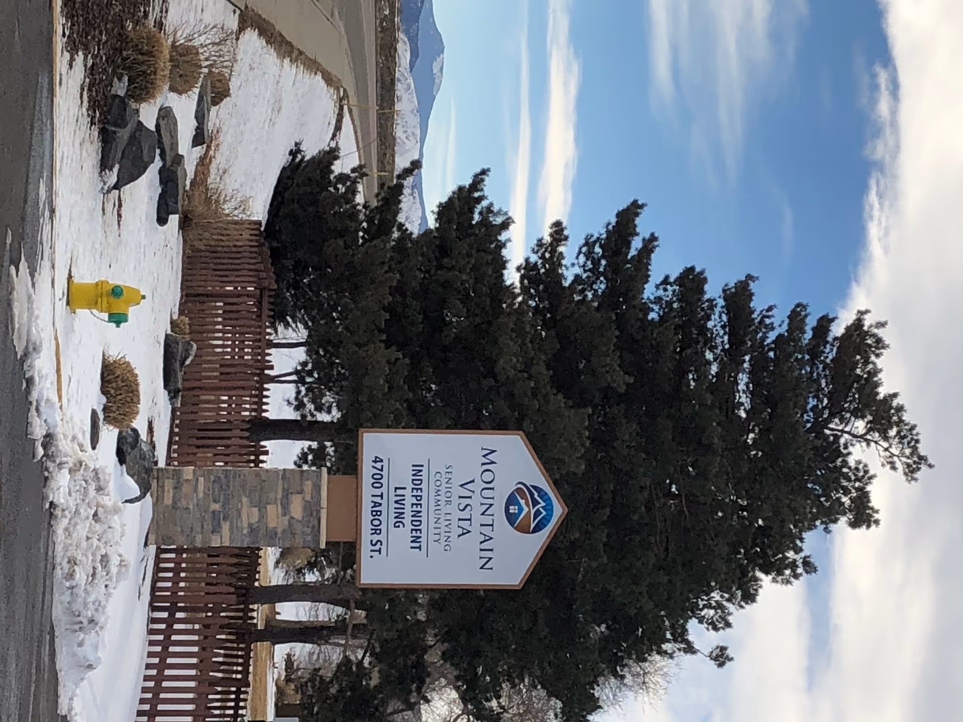 Entrance sign for Mountain Vista Senior Living Community beside a stone pillar, evergreen trees, and snow-covered ground.