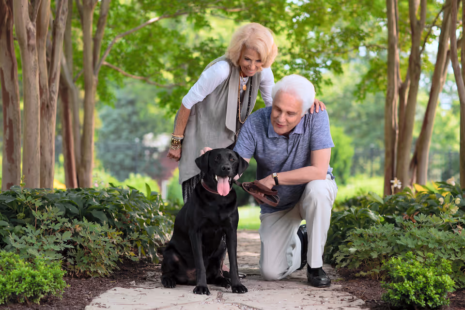 An elderly couple outdoors in a garden-like setting with trees and greenery. The woman is standing and smiling while the man is kneeling beside a black dog with its tongue out. Both appear happy and engaged with the dog.