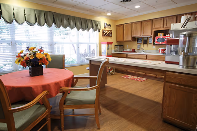 A cozy kitchen and dining area with wooden cabinets, a countertop, and a round table covered with a red tablecloth. The table has a floral centerpiece and is surrounded by wooden chairs with green cushions. Large windows with green valances let in natural light.