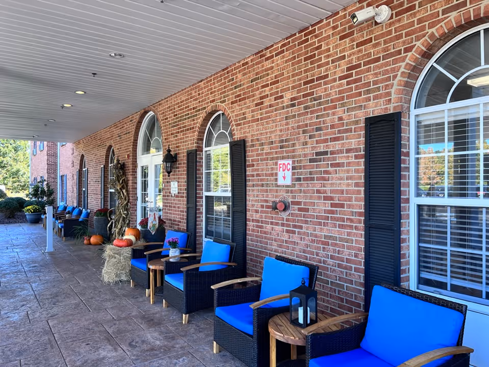 Covered brick building entrance with a row of blue-cushioned chairs, small tables and fall decorations including pumpkins and hay bales.
