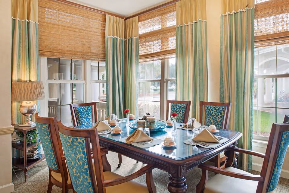 Sunlit dining room with a wooden table set for six, upholstered chairs, and large windows with bamboo shades and green curtains.