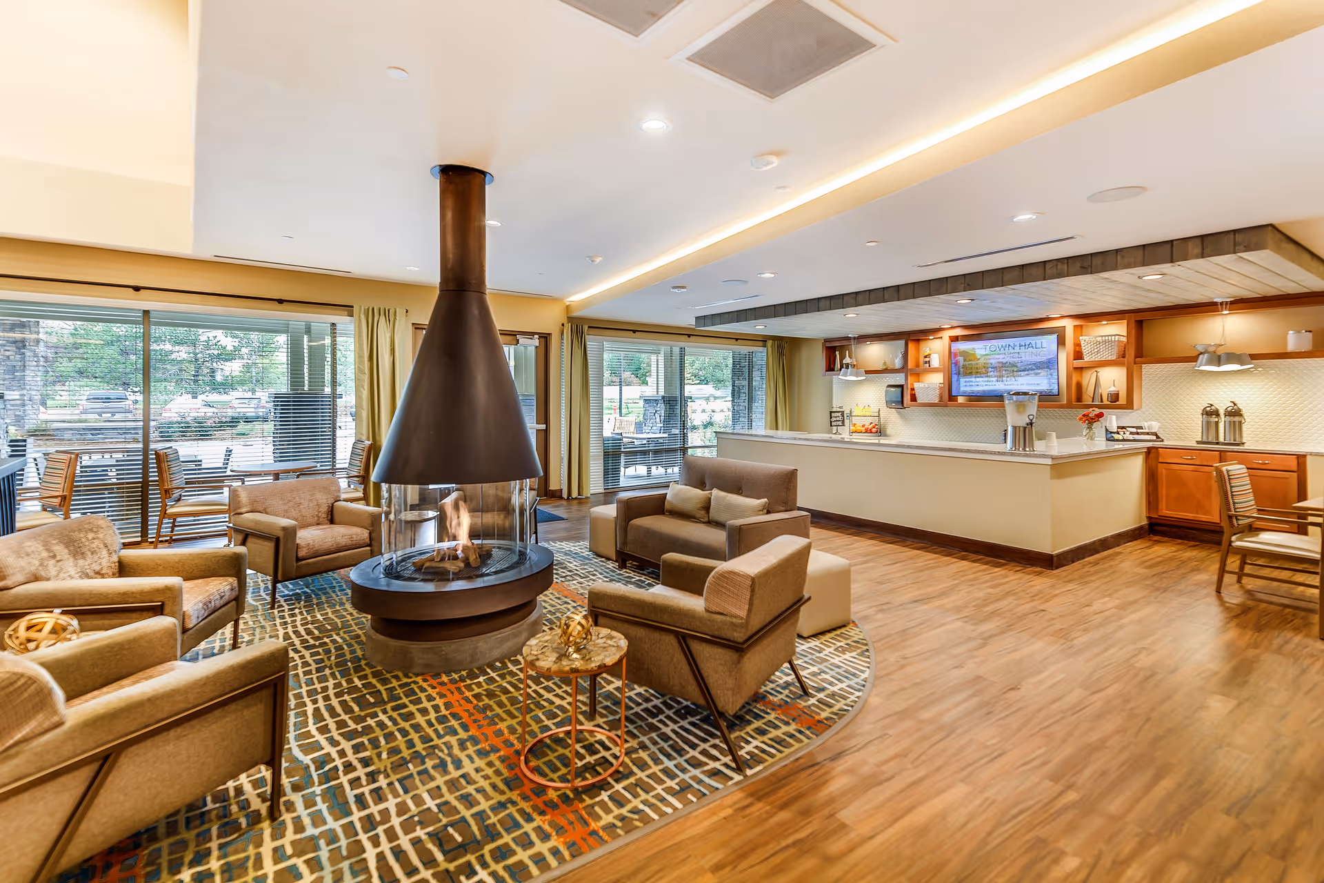 A bright communal lounge with a central suspended fireplace surrounded by armchairs, large windows, and a service counter area.