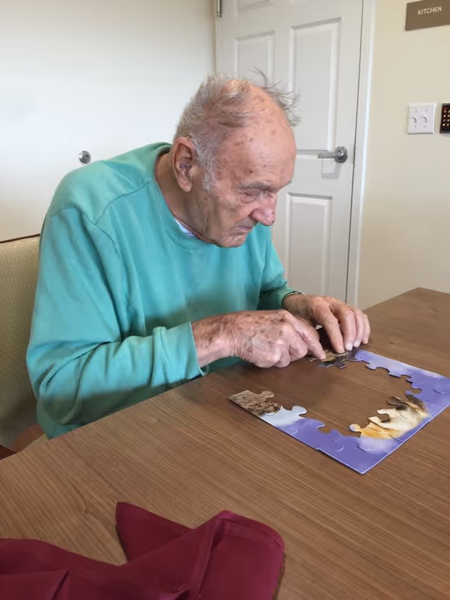 An elderly man wearing a green sweatshirt is sitting at a wooden table working on assembling a jigsaw puzzle. Behind him is a white door with a sign labeled 'KITCHEN' on the wall next to it.