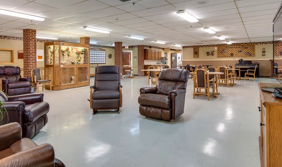 A spacious common area in a senior living facility featuring several brown leather recliners arranged in the foreground and multiple wooden tables with chairs in the background. The room has a tiled ceiling with fluorescent lighting, brick pillars, and a piano against the far wall. There is also a wooden display case with decorative flowers and a small kitchen area visible in the back.