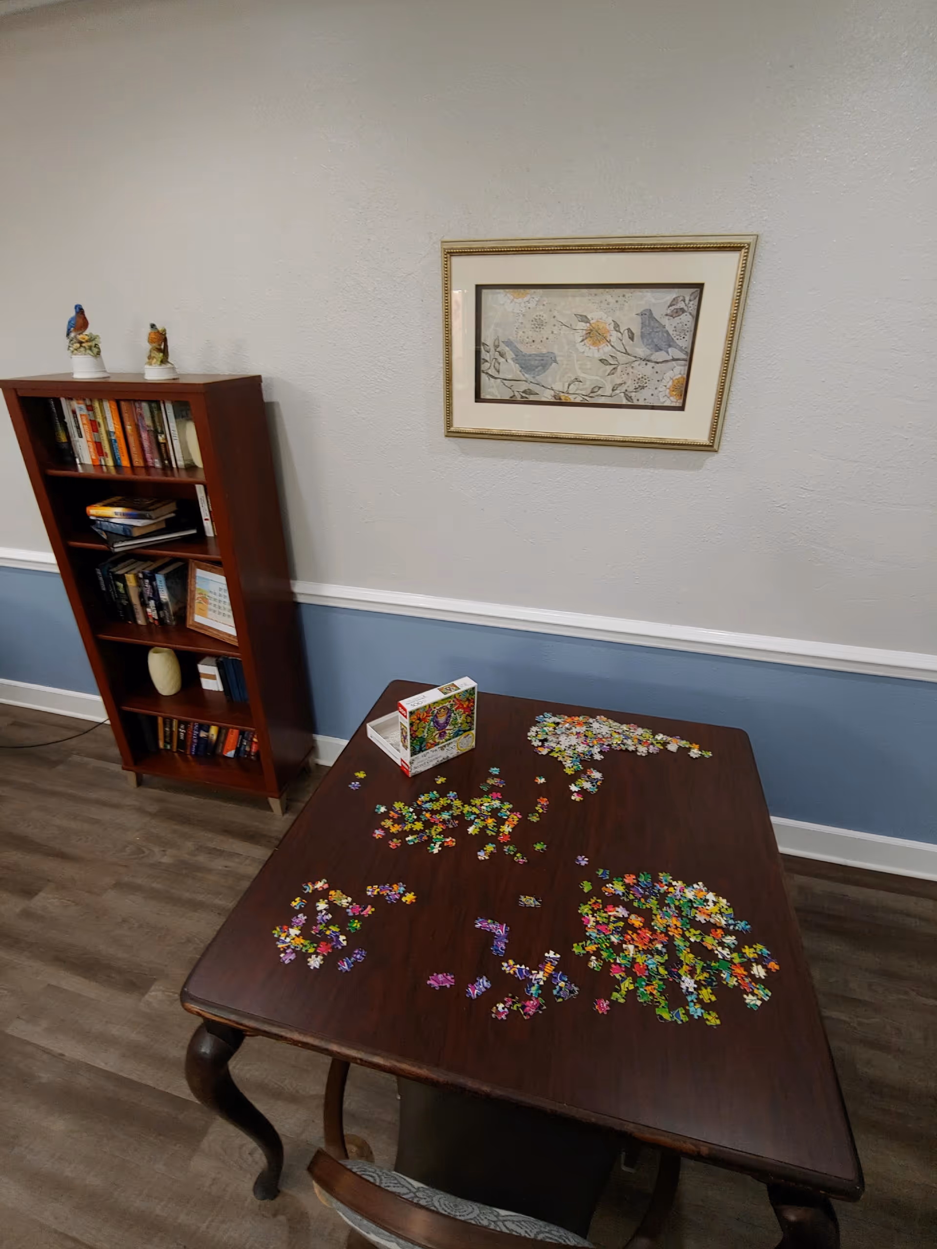 A wooden table with an incomplete colorful jigsaw puzzle spread out on it, located in a room with blue and white walls. A wooden bookshelf filled with books and decorative bird figurines is against the wall, and a framed picture of birds and flowers hangs above the table.