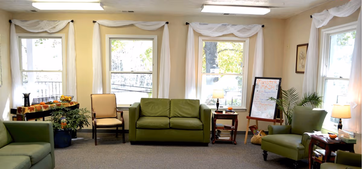 A bright living room with three large windows draped with white curtains. The room features green upholstered sofas and armchairs, a beige armchair, two small wooden side tables with lamps, a potted plant, and a table with a fall-themed tablecloth holding a bowl of fruit. There is also a whiteboard with colorful writing and a basket on the floor.