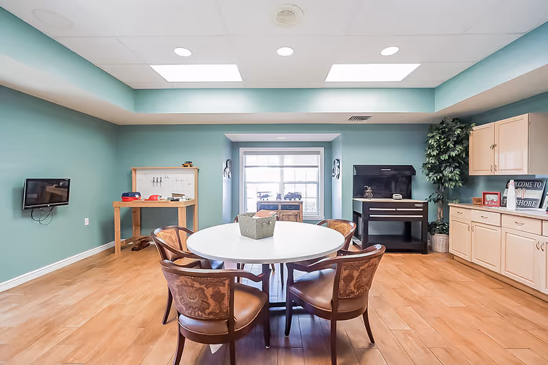 Bright communal activity room with a round table and six chairs, teal walls, cabinets and a wall-mounted TV.