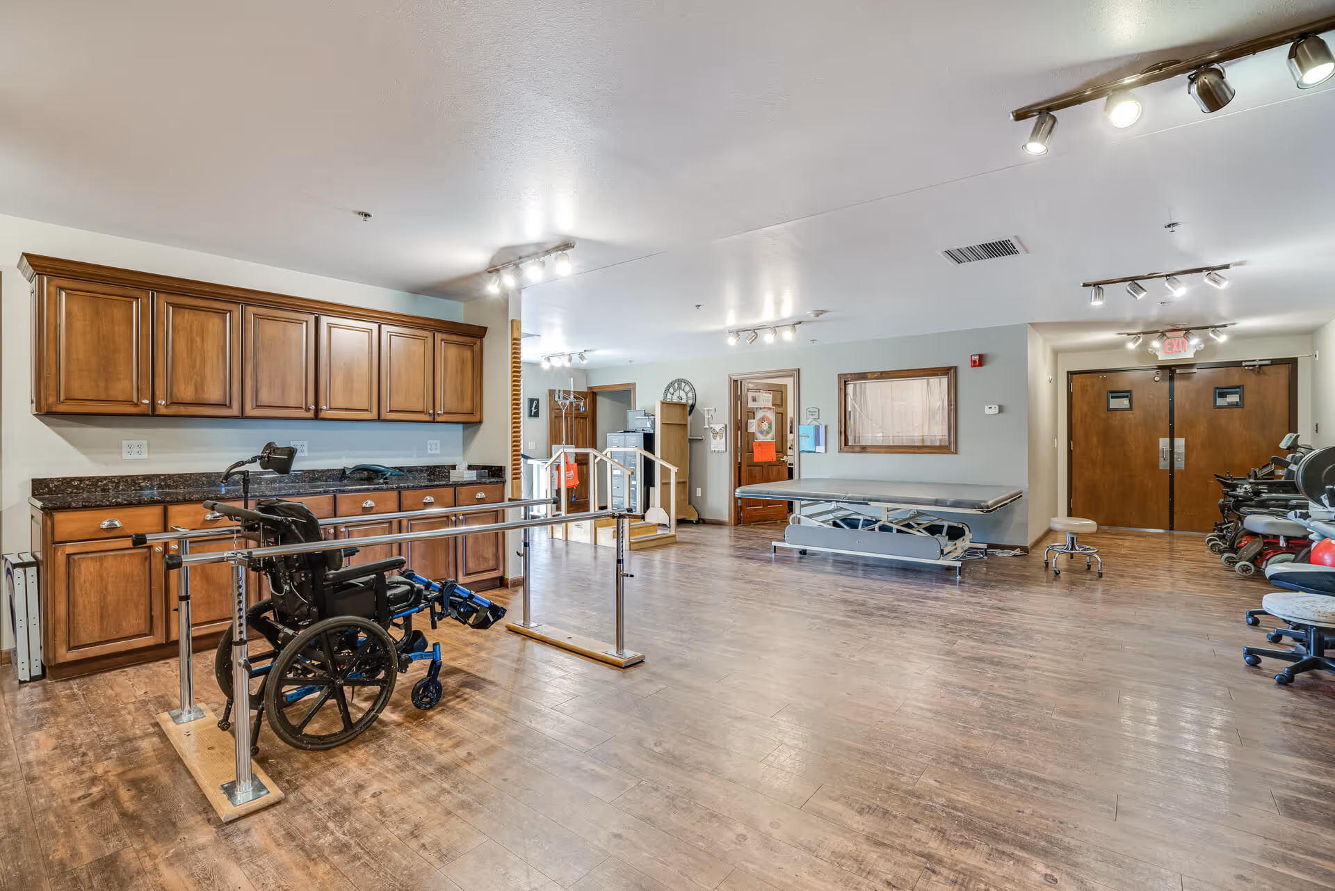 A spacious rehabilitation room with parallel bars, a wheelchair, treatment tables, and wooden cabinetry in a senior living facility.