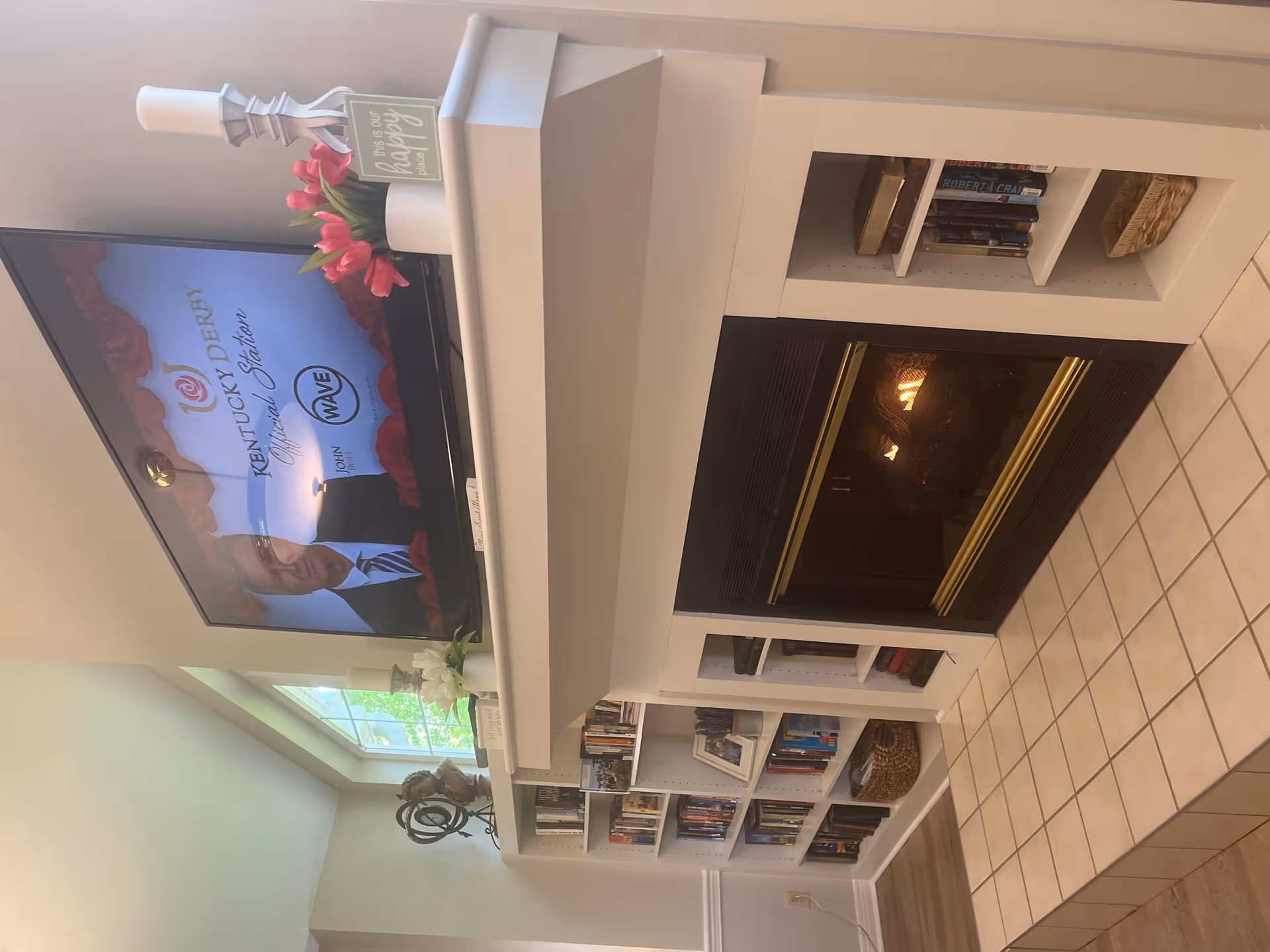 A cozy living room area featuring a lit fireplace with a white mantel. Above the mantel is a flat-screen TV displaying a Kentucky Derby official station program. On the mantel are decorative items including a candle holder, a small sign that reads 'this is our happy place,' and a vase with pink flowers. Built-in white bookshelves flank the fireplace, filled with books and decorative items. A large window allows natural light to brighten the space.