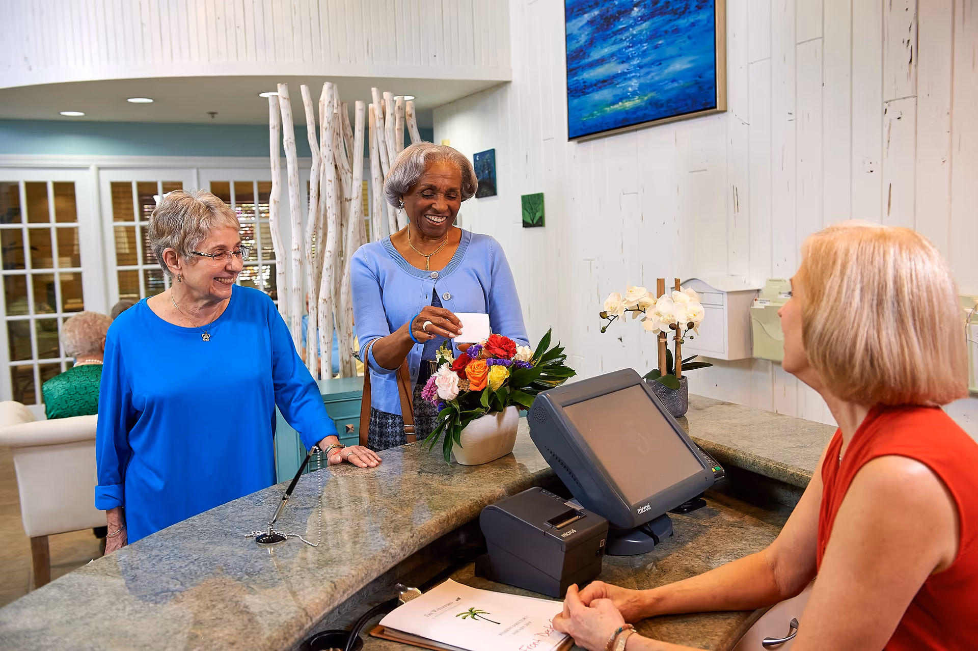 Two elderly women standing at a reception desk in a senior living facility, one woman holding a card and smiling while the receptionist, a blonde woman in a red sleeveless top, looks on. The reception area has a granite countertop, a computer, a vase with colorful flowers, and white wooden walls with artwork.