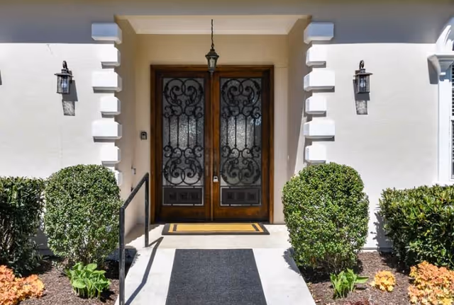 Entrance to a building with double wooden doors featuring decorative wrought iron glass panels. The doors are framed by white walls with decorative trim, two wall-mounted lantern-style lights on either side, and neatly trimmed bushes and plants along the walkway leading to the entrance.