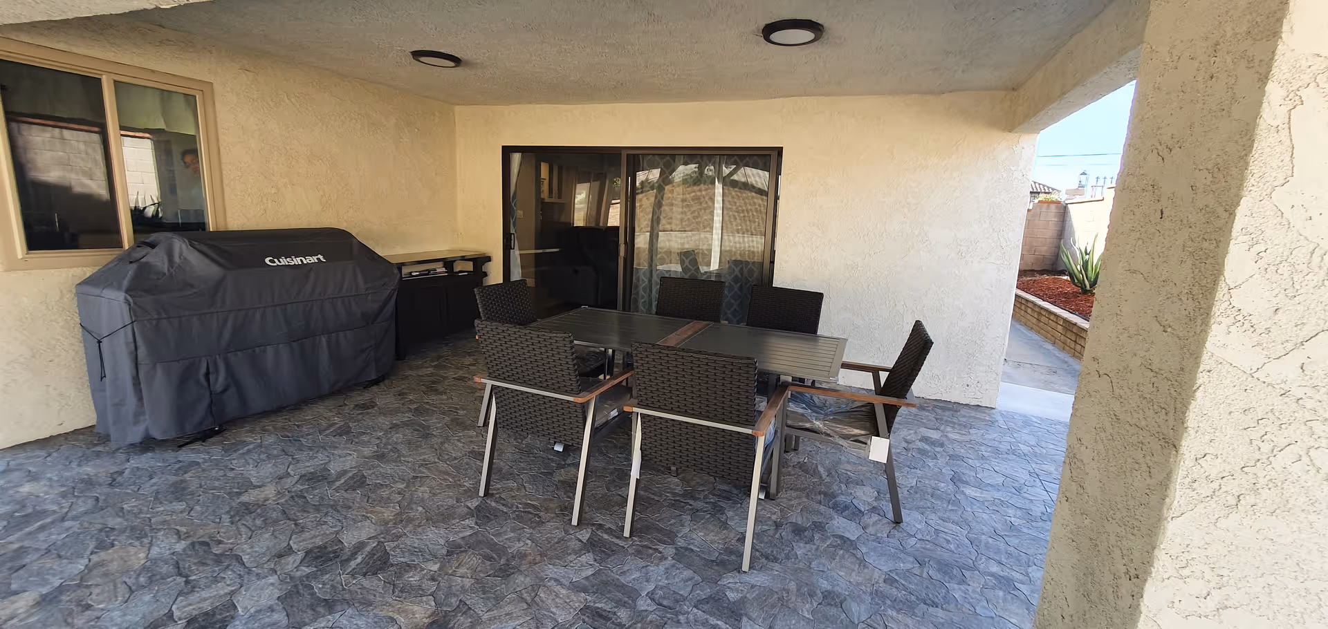 Covered outdoor patio area with a stone-patterned floor, a black dining table with six wicker chairs, a covered Cuisinart grill, and sliding glass doors leading inside. The patio is surrounded by beige stucco walls and has a small garden bed visible on the right side.