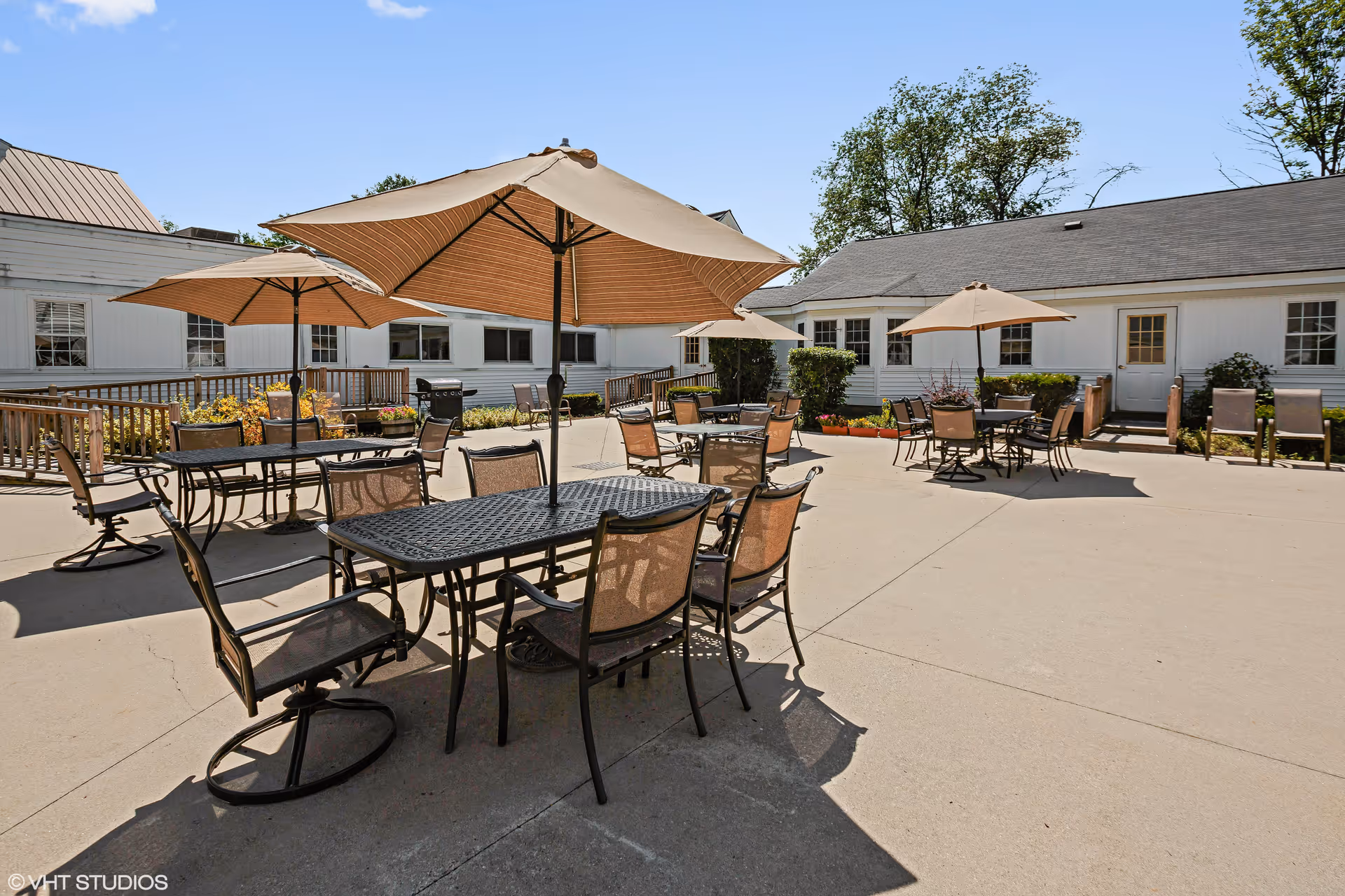 Outdoor patio area with multiple tables and chairs, each table shaded by large beige umbrellas. The patio is surrounded by white buildings with windows and doors, and there are some plants and bushes along the edges. The sky is clear and blue.