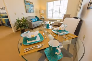 A bright and cozy senior living apartment interior featuring a glass dining table set for two with turquoise and white dishware, a small living area with a gray loveseat, a side table with a lamp, a large window letting in natural light, and a glimpse of a bedroom in the background.
