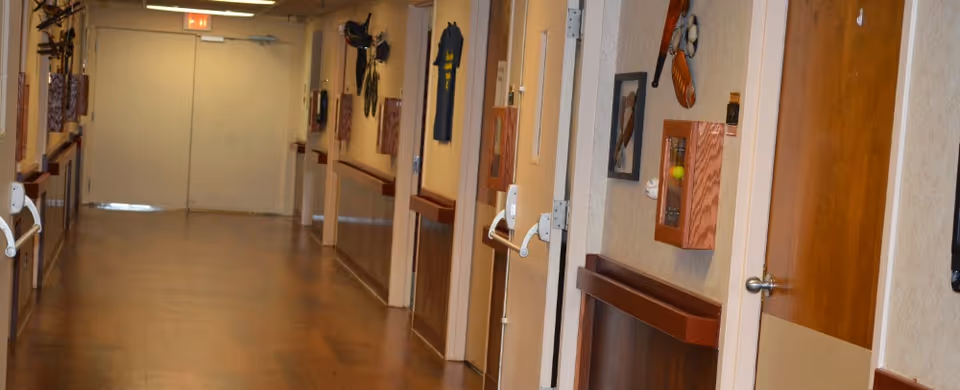 A long indoor hallway with wooden floors and beige walls, decorated with various sports memorabilia such as baseball gloves, bats, and framed pictures. Several doors line the hallway, and handrails are mounted on the walls. An exit sign is visible above double doors at the end of the hallway.