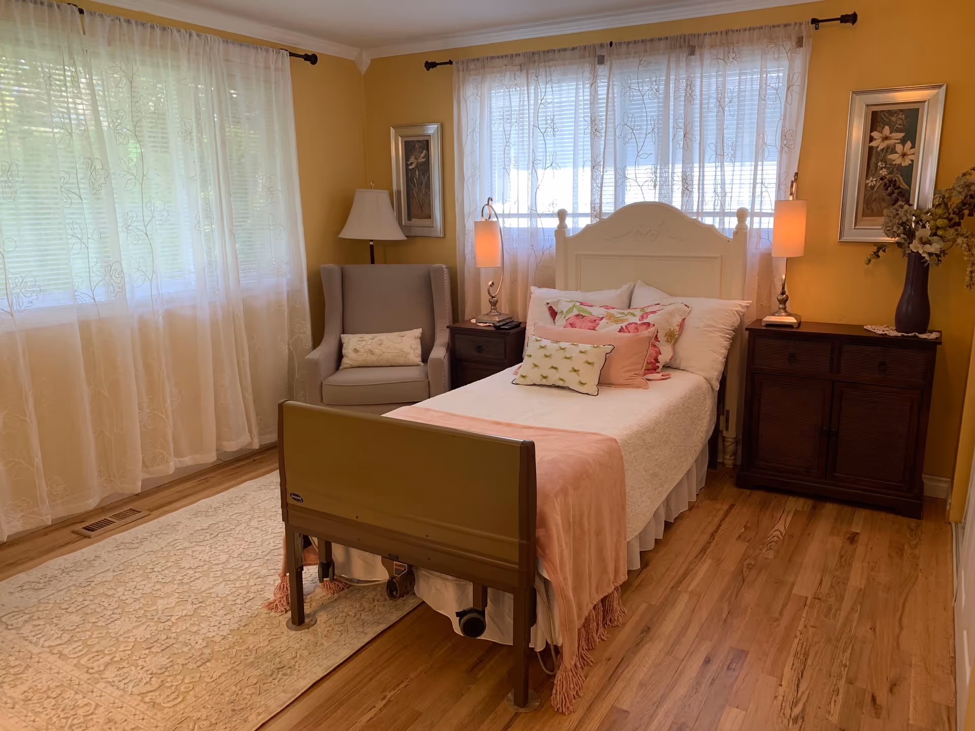 A cozy bedroom with a single bed featuring white and pink bedding, a wooden headboard, and a footboard. The room has yellow walls, wooden flooring, and sheer white curtains covering two windows. There is a gray upholstered armchair with a decorative pillow next to a floor lamp on the left side of the bed. On the right side, there is a dark wooden dresser with a vase of flowers and two framed floral pictures on the walls. Two bedside tables with lamps flank the bed.