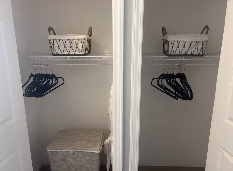Two open closet spaces with white walls and white wire shelves. Each closet has a black wire basket with white fabric lining on the top shelf and several black hangers hanging from the rod below. The left closet also contains a beige woven hamper with a lid on the floor.