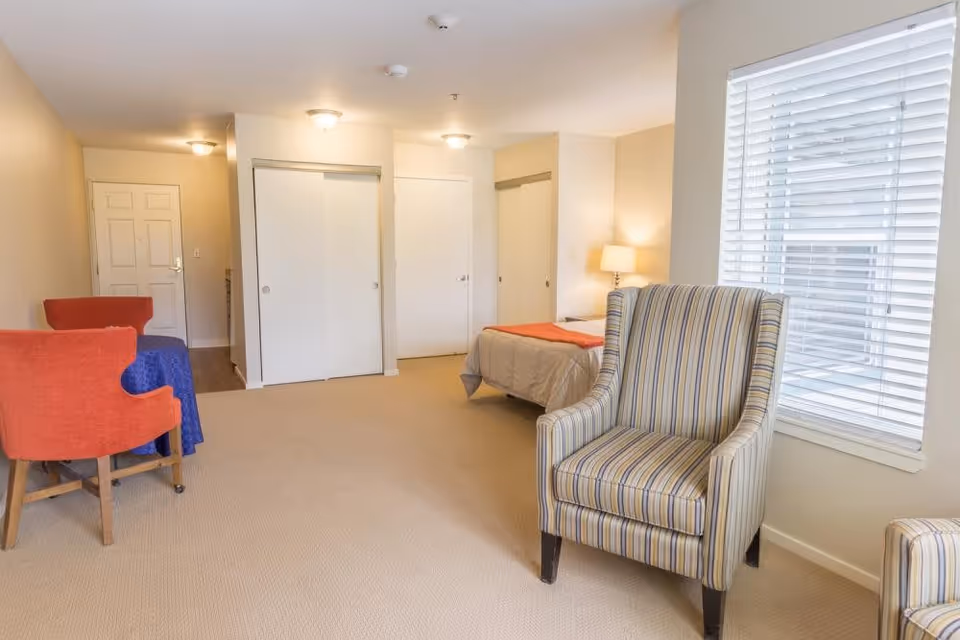 A bright, spacious senior living bedroom with beige walls and carpet. The room features a bed with a beige comforter and an orange throw, a striped armchair near a window with white blinds, and two orange chairs around a small table covered with a blue cloth. There are multiple white doors and closets along the back wall, and ceiling lights provide illumination.