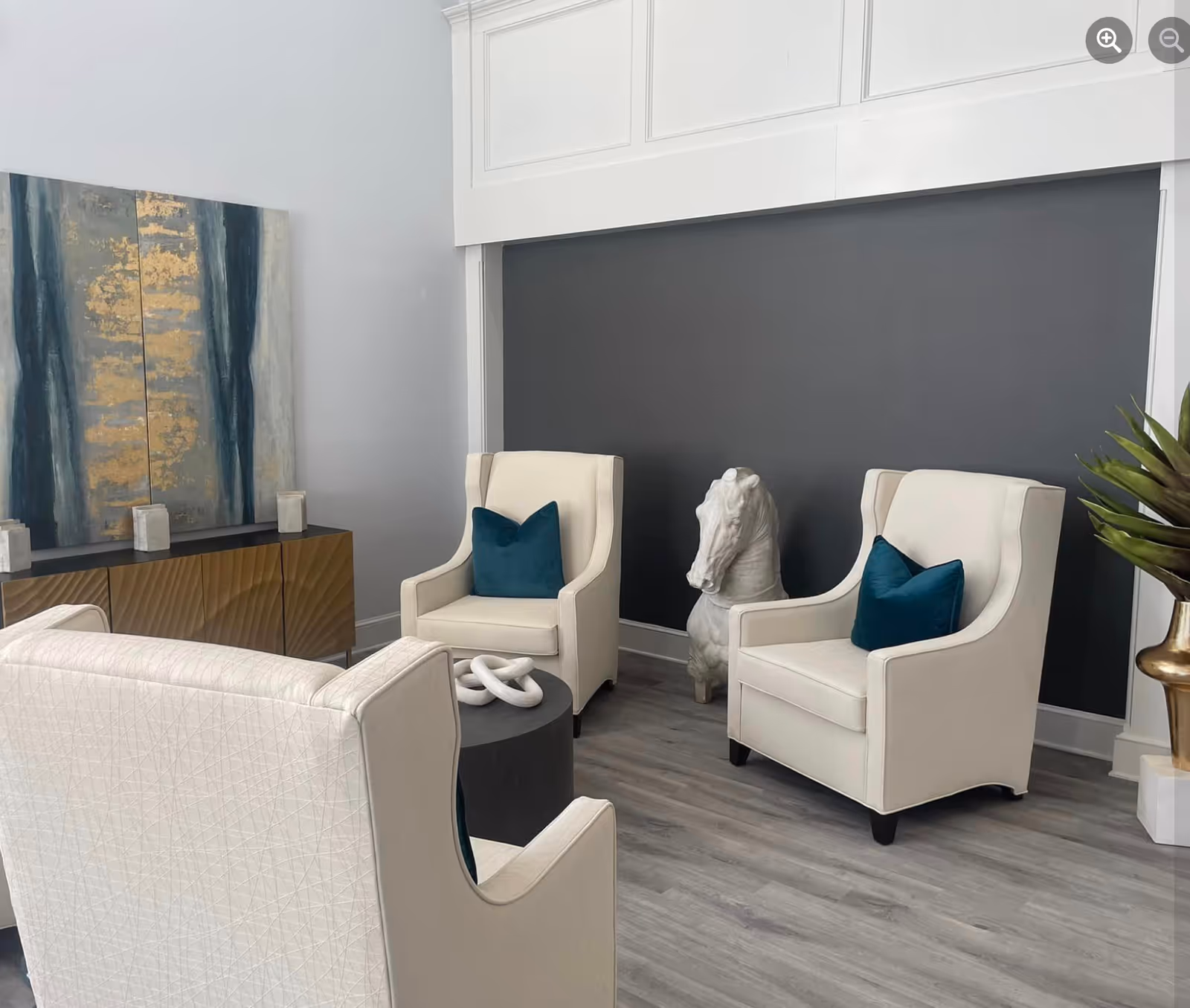 A modern sitting area with three cream-colored armchairs, each with a dark teal pillow. There is a small round black table with a white decorative object on it. Behind the chairs is a dark gray accent wall with a white horse head sculpture on the floor. To the left, there is a wooden cabinet with a textured front and a large abstract painting above it. The floor is light gray wood, and a green plant in a gold vase is partially visible on the right.