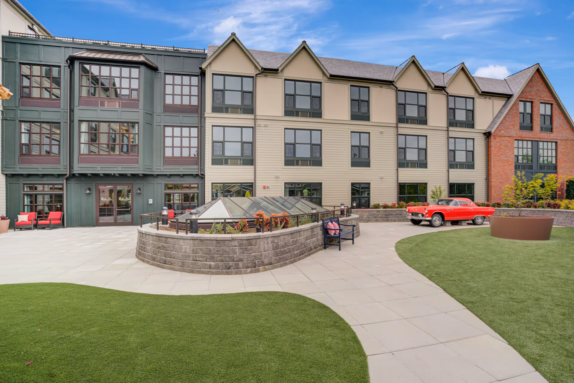 Outdoor courtyard area of a senior living facility with a paved walkway, green lawn, a raised planter with plants, a bench with a cushion, and a vintage red car parked near the building. The building has multiple large windows and a mix of green, beige, and red brick exterior walls under a blue sky with some clouds.