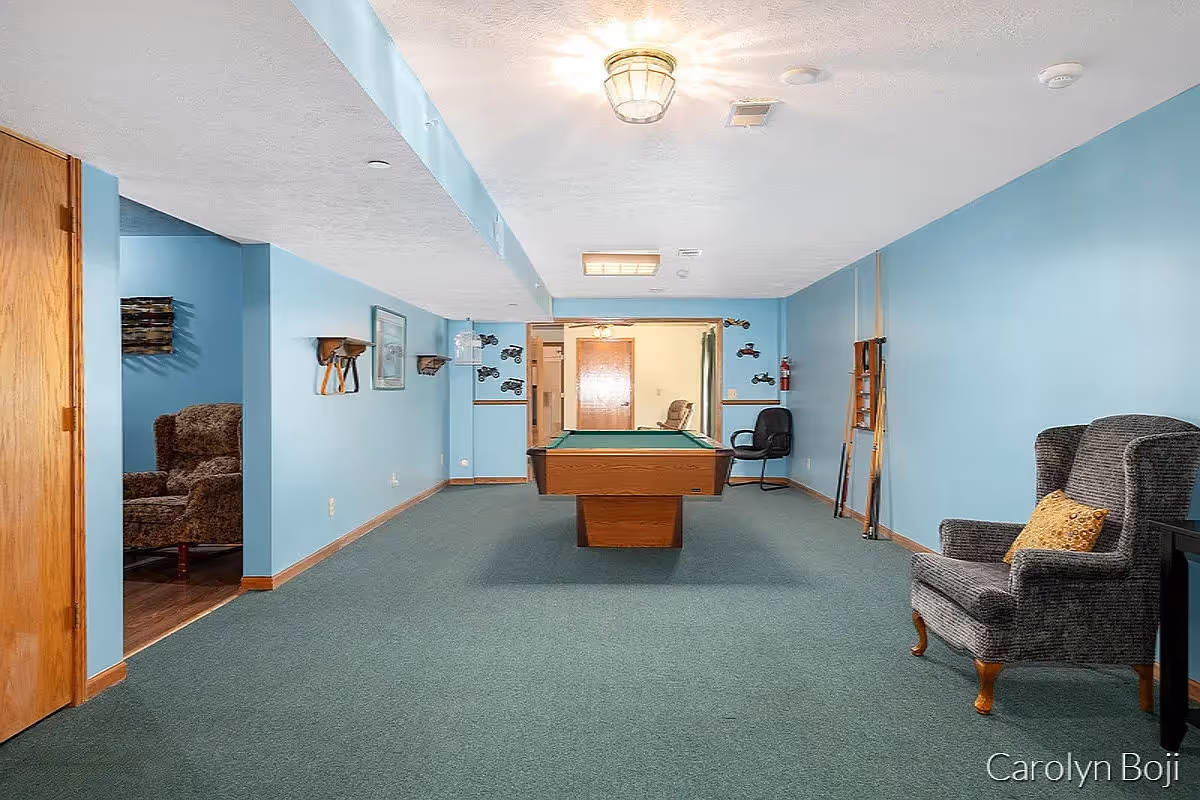 A recreation room with blue walls and green carpet featuring a pool table in the center. There are two armchairs, one gray with a yellow pillow on the right and a brown patterned one partially visible in an adjacent room on the left. Pool cues are mounted on the right wall, and there are small decorative shelves and framed pictures on the left wall. The room is lit by ceiling lights.