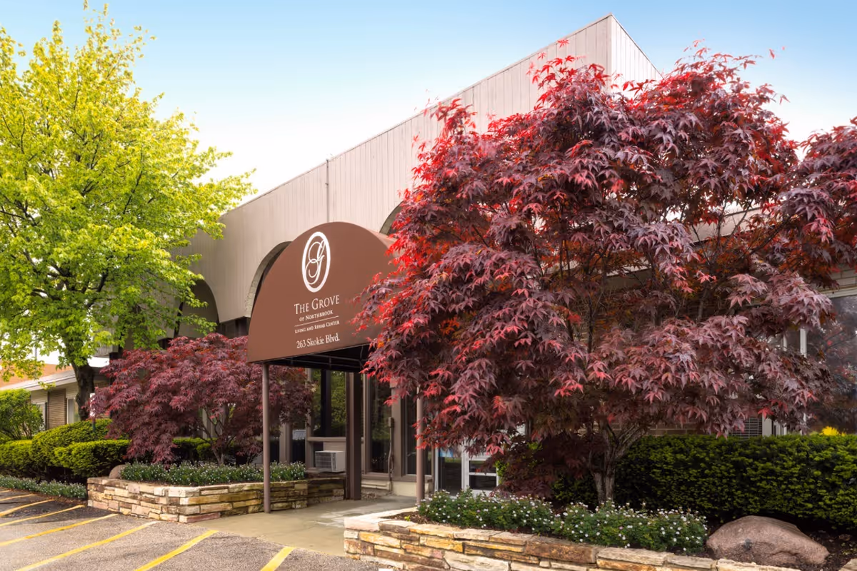 Front entrance of The Grove of Northbrook with a brown canopy, stone planters, and surrounding trees and landscaping.