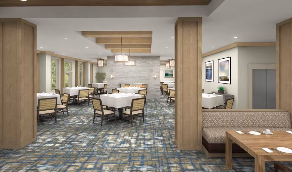 A spacious dining room with multiple tables covered in white tablecloths and surrounded by wooden chairs with cushioned seats. The room features large windows letting in natural light, a patterned carpet, wooden ceiling beams, and a modern fireplace on the far wall. There is also a booth seating area with a wooden table set with plates and cups.